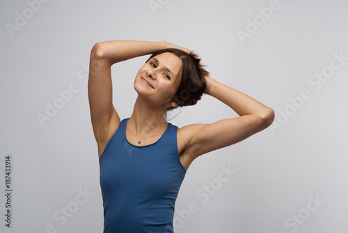 Slim attractive girl with beautiful tanned skin and athletic physique smiling and looking at camera Posing holding her dark straight hair gathered on head. Portrait in blue shirt on white background.