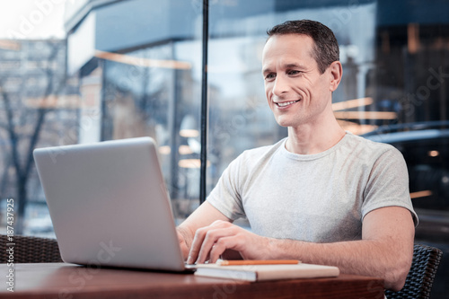 Modern technologies. Cheerful brunette keeping smile on his face and holding hands on keyboard while looking at screen of his computer