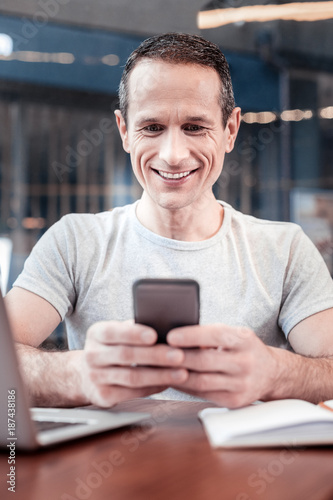 Joyful mood. Positive delighted male person sitting at the table and looking at his gadget while communicating online