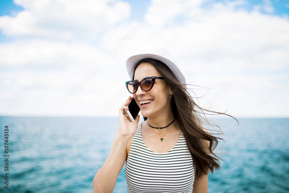 Beautiful young modern brunette woman in sunglasses talking on the phone with her friend on the beach against the sea