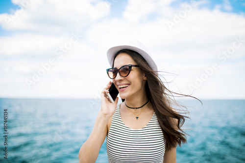 Beautiful young modern brunette woman in sunglasses talking on the phone with her friend on the beach against the sea