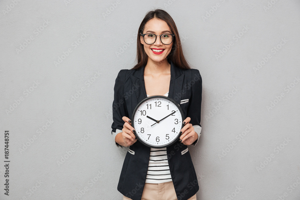 Smiling woman in eyeglasses holding clock and looking at camera Smiling woman in eyeglasses holding clock and looking at camera