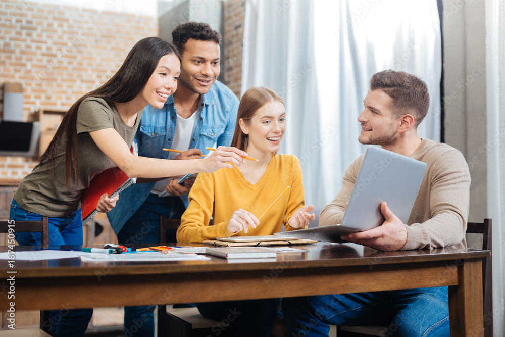 Amazing. Young smiling student holding a modern laptop while his positive friends looking attentively at the screen of it and feeling surprised Amazing. Young smiling student holding a modern laptop while his positive friends looking attentively at the screen of it and feeling surprised