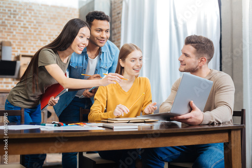 Amazing. Young smiling student holding a modern laptop while his positive friends looking attentively at the screen of it and feeling surprised