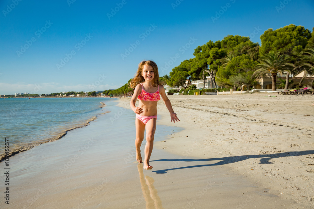 Cute happy little girl running along the beach in swimming suit, jumping over waves. Beautiful summer sunny day, blue sea, turquoise water, picturesque landscape. Majorca, Spain Cute happy little girl running along the beach in swimming suit, jumping over waves. Beautiful summer sunny day, blue sea, turquoise water, picturesque landscape. Majorca, Spain