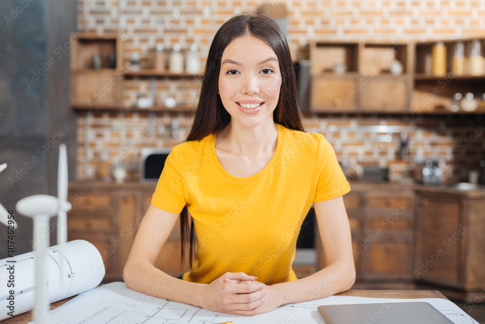 Diligent employee. Cheerful emotional young engineer looking friendly while sitting at the table with schemes and drawings in front of her and smiling kindly Diligent employee. Cheerful emotional young engineer looking friendly while sitting at the table with schemes and drawings in front of her and smiling kindly