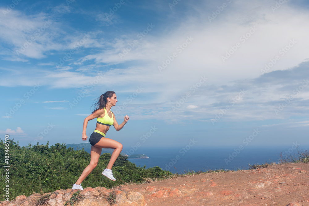 Athletic woman running up the mountain with sky and sea in background. Professional runner doing cardio work-out outdoor in natural landscape Athletic woman running up the mountain with sky and sea in background. Professional runner doing cardio work-out outdoor in natural landscape