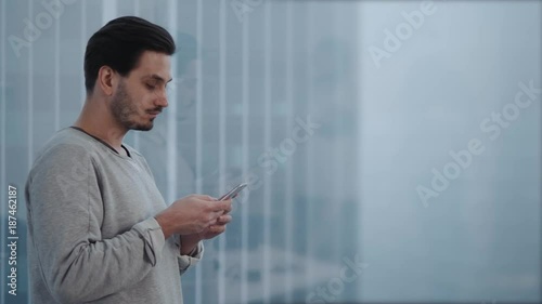 Portrait of young businessman talk on cellphone while stand by his office window in modern interior of skyscraper building, male entrepreneur having mobile phone conversation after important briefing photo