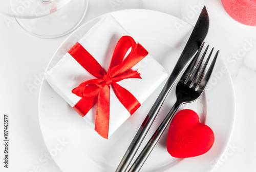 Valentine's day table setting with plate, fork, knife, gift box and red heart, on white marble background copy space top view