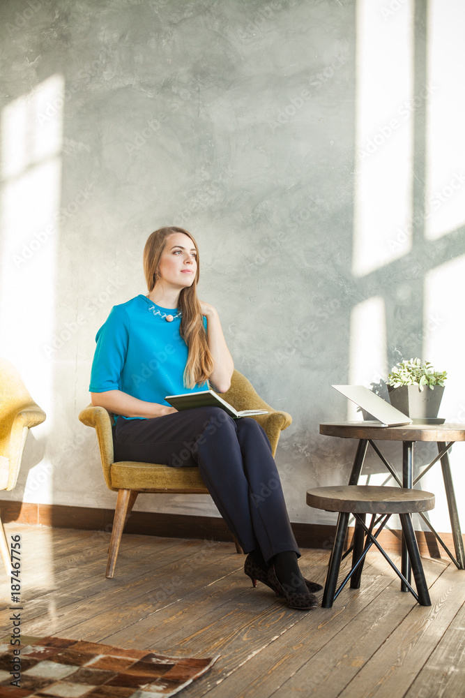 Fototapeta premium Cheerful Woman with Book and Laptop at Home Office. Woman Working with New Startup Project in Modern Loft