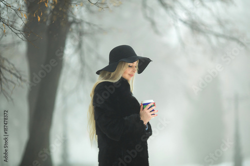 beautiful young girl in fur coat and hat walking in the park in winter weather with a glass of coffee in hands © Alexandr