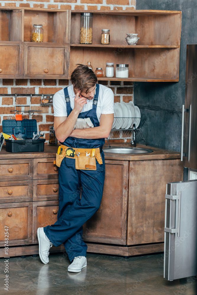 pensive young repairman looking at broken refrigerator in kitchen - obrazy, fototapety, plakaty 