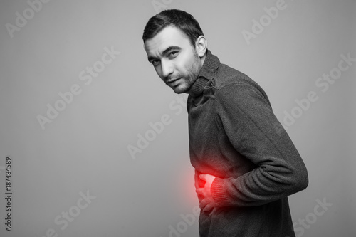 young man holding his sick stomach in pain, isolated on gray background, monochrome photo with red as a symbol for the hardening photo