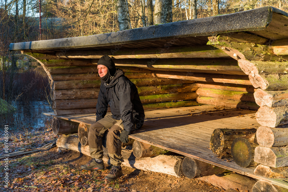 Male hiker sitting in wind shelter, relaxing and warming up in the morning sunshine. Ice on the river behind the shelter.