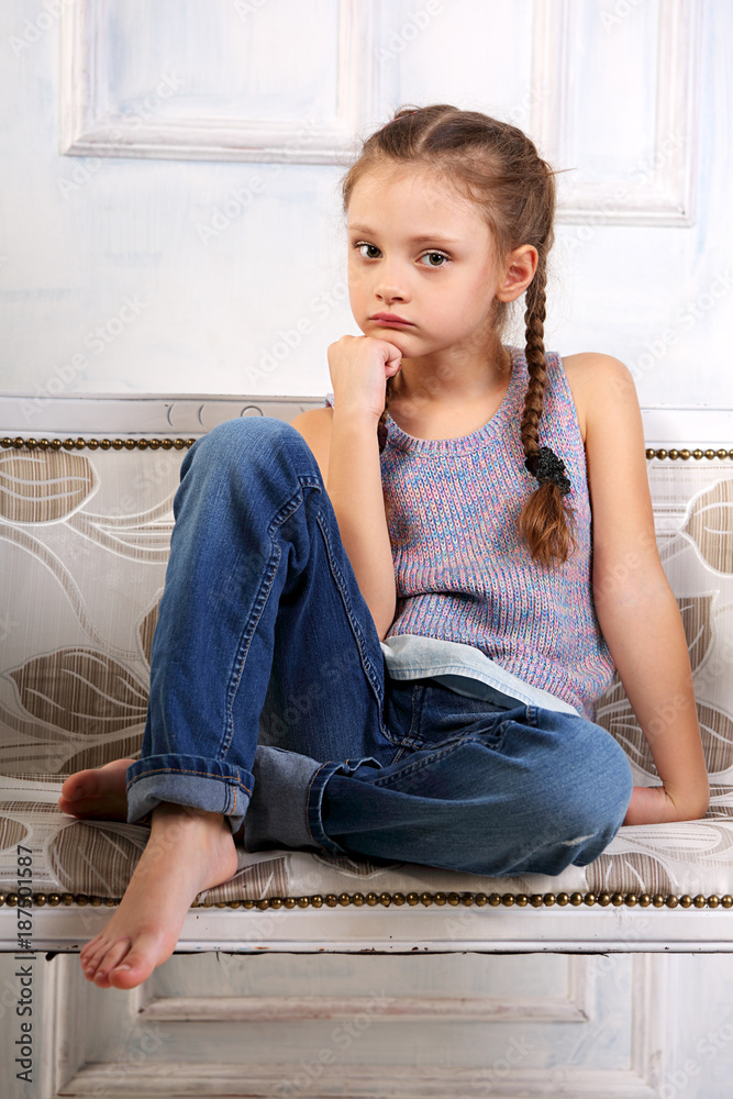 Beautiful calm thinking unhappy kid girl sitting on the bench in blue jeans  and fashion blouse looking happy. studio portrait Stock Photo | Adobe Stock