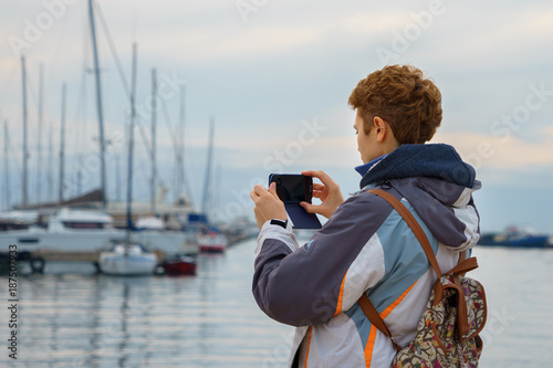 A tourist girl with a backpack is standing on the waterfront in the marina and is going to take photo of yachts and motor boats on her mobile phone