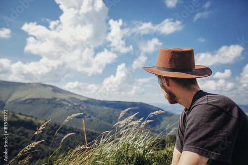 wanderlust and travel concept. stylish traveler man in hat looking at mountains. hipster guy traveling on top of mountain, relaxing at windy grass. space for text. atmospheric moment © sonyachny