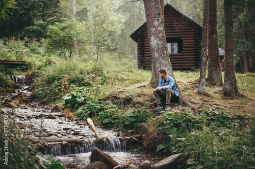 Leinwand Poster stylish hipster man sitting and relaxing at wooden cabin in forest