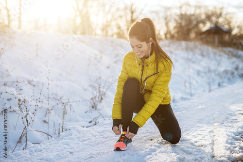 Portrait view of young satisfied smiling motivated and focused sporty active girl with a ponytail crouching in winter sportswear the snowy nature on the road and tying shoelaces on sneakers.