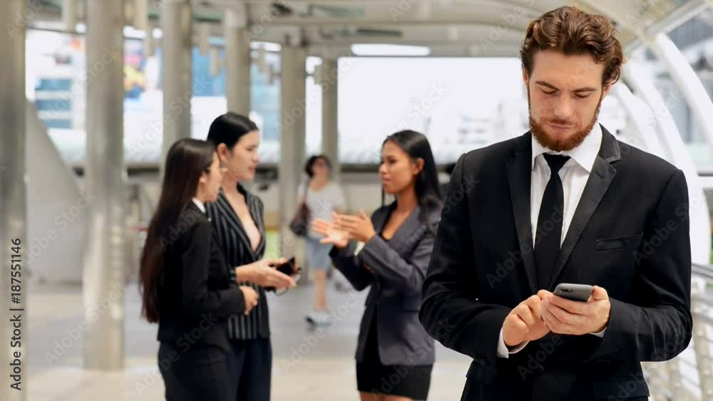 Young business man having a private phone conversation during his break outside office with his friends in background talking. Trying to close the deal. Medium shot. Mobile office concept. Young business man having a private phone conversation during his break outside office with his friends in background talking. Trying to close the deal. Medium shot. Mobile office concept.
