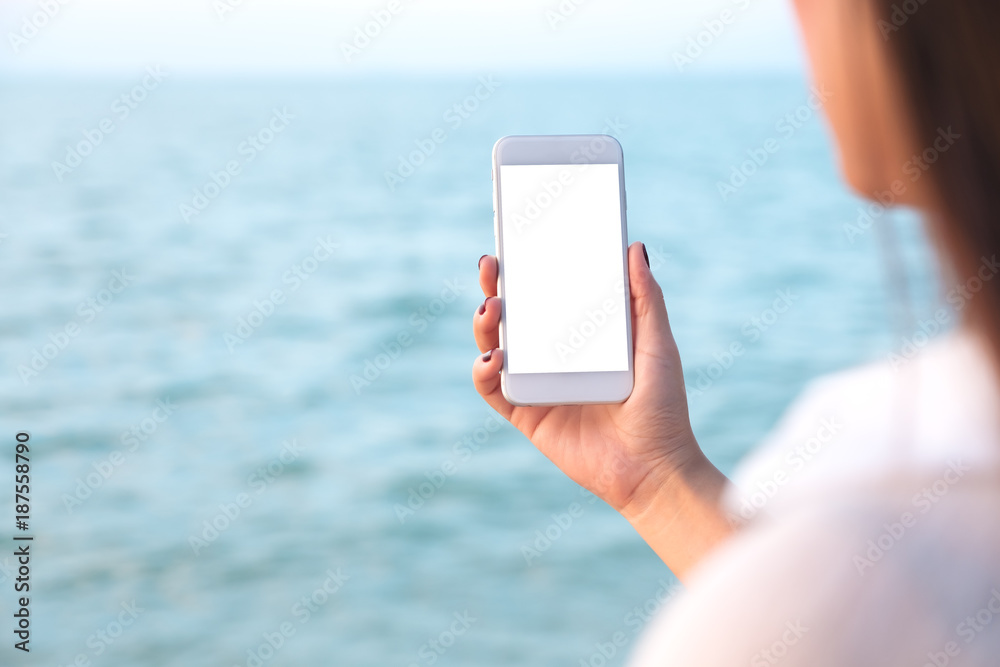 Mockup image of a woman's hand holding white mobile phone with blank desktop screen by the sea and blue sky background