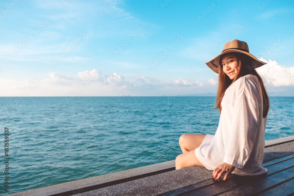 Portrait image of a happy beautiful asian woman on white dress sitting on wooden balcony by the sea with clear blue sky background