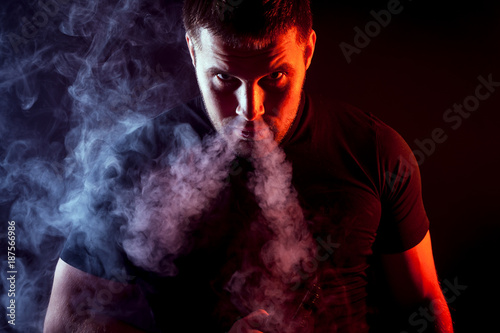 A young stylish male smoker in a black T-shirt stares and exhales from the nose a large cloud of smoke from the vape on a dark isolated background