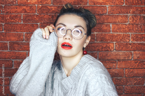 Wonderful portrait of cute charming girl with beautiful eyes and round glasses with her mouth slightly open in a white cardigan on brick background, closeup. photo