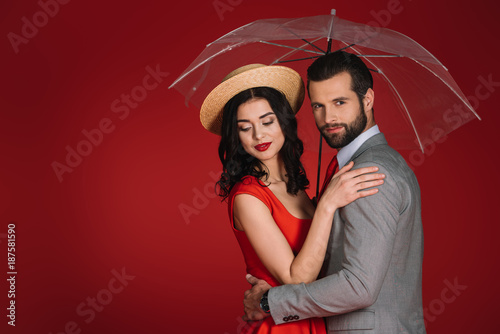 attractive couple hugging under transparent umbrella isolated on red