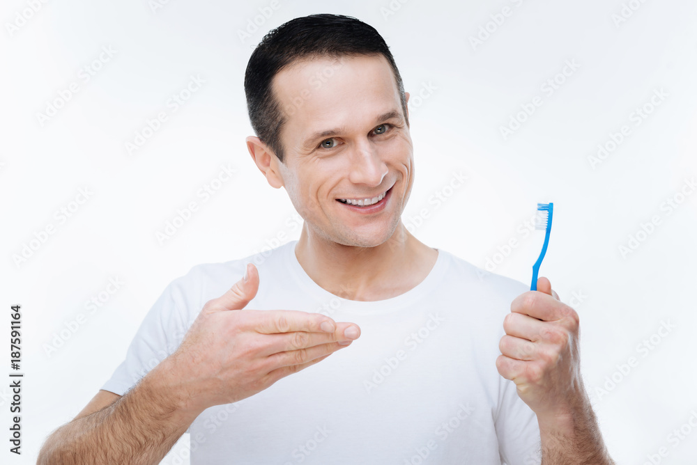 Dental care. Positive cheerful nice man pointing at his toothbrush and smiling while caring about his teeth Dental care. Positive cheerful nice man pointing at his toothbrush and smiling while caring about his teeth
