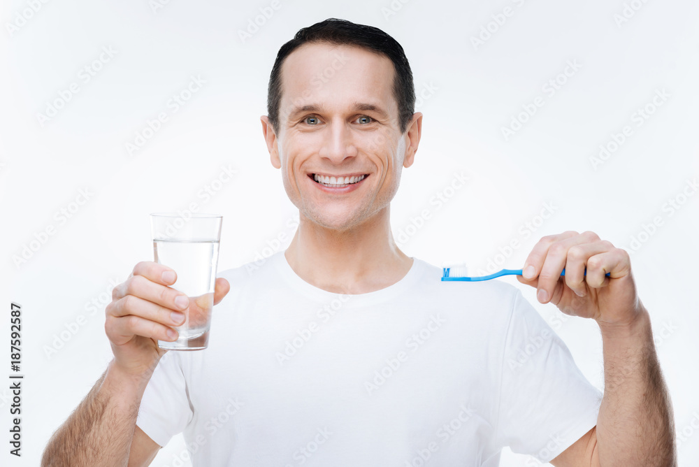 Teeth hygiene. Nice happy delighted man holding water and a toothbrush and smiling while looking at you Teeth hygiene. Nice happy delighted man holding water and a toothbrush and smiling while looking at you