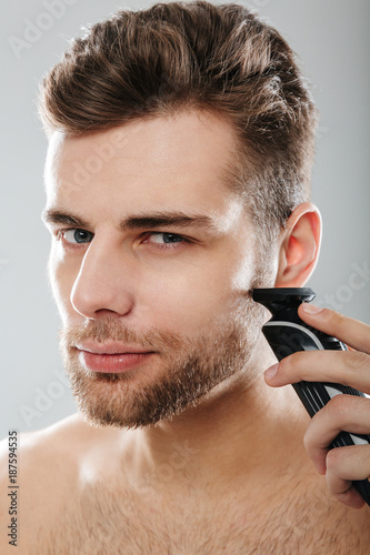 Closeup picture of good-looking adult guy grooming his face with shaving his cheek using electric shaver over grey wall