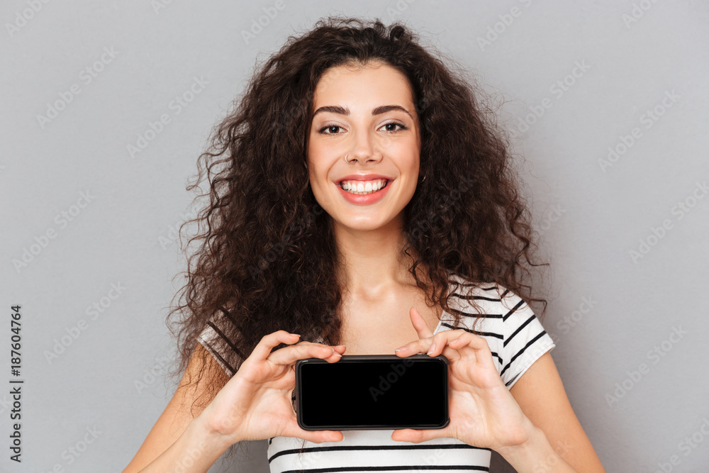 Fototapeta premium Optimistic woman with ring in nose holding her mobile phone meaning good choice smiling on camera while isolated against grey wall close up
