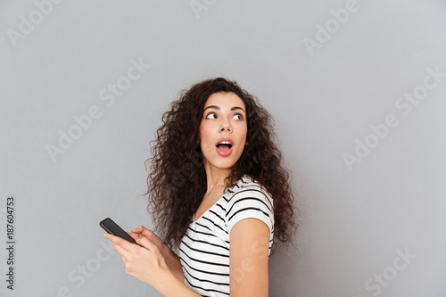 Curious brunette woman with mobile phone in hand turning around with open mouth like watching something interesting or breathtaking