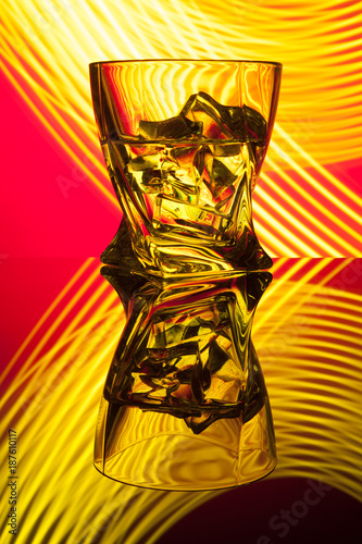 Cocktail whiskey a glass with pieces ice of party reflection a concept of hourglasses yellow light effects on red background.