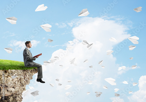 Man on edge reading book and paper planes flying in air