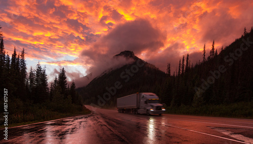 Truck on highway with reflections  in Canada surrounded by mountains in clouds during orange sunset © Martin Hossa