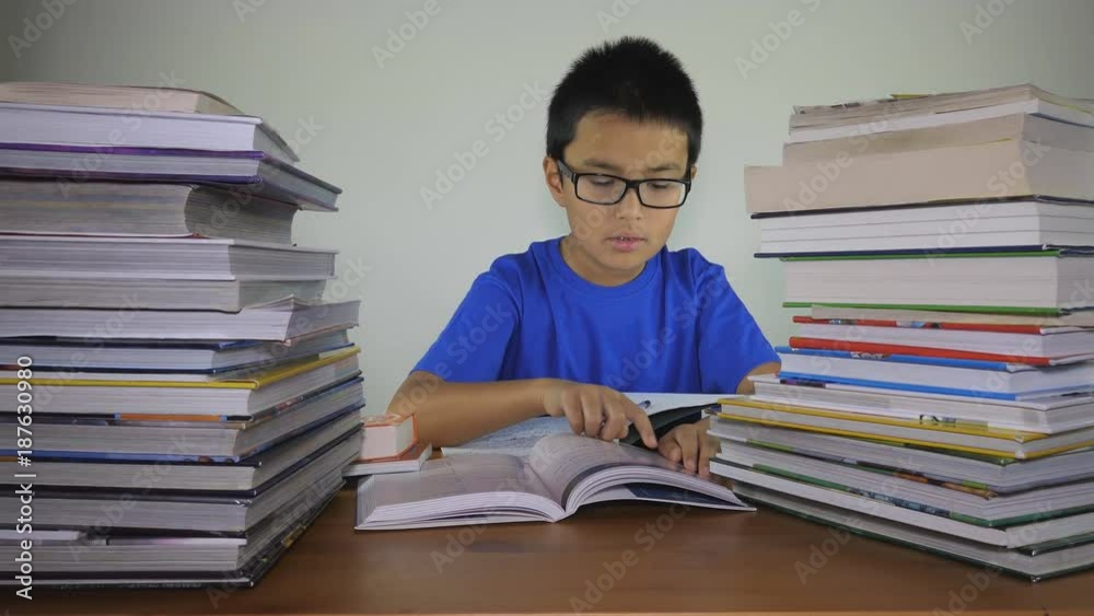 A boy is doing his homework on a table with stacks of books on both sides. A boy is doing his homework on a table with stacks of books on both sides.