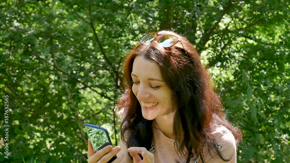 Amazing Girl Laughs Reading Message in Social Network on Her Mobile Phone. Closeup Portrait of Beautiful Brunette Woman with Blue Smartphone Outdoors on Green Trees Background. Amazing Girl Laughs Reading Message in Social Network on Her Mobile Phone. Closeup Portrait of Beautiful Brunette Woman with Blue Smartphone Outdoors on Green Trees Background.
