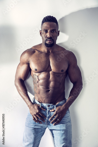 African American bodybuilder man, naked muscular torso, wearing jeans, leaning on white wall