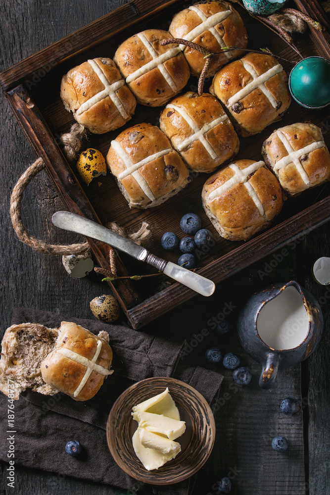 Fototapeta premium Hot cross buns in wooden tray served with butter, knife, blueberries, easter eggs, birch branch, jug of cream on textile napkin over old texture wood background. Top view, space. Easter baking.