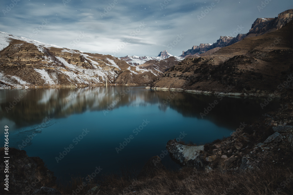 High mountain lake in the northern Caucasus, surrounded by epic rocks and a bright winter starry sky at sunrise.