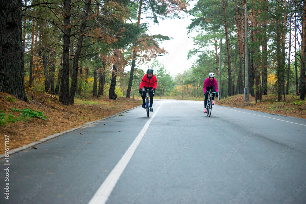 Two Young Female Cyclists Riding Road Bicycles in the Park in the Cold Autumn Morning. Healthy Lifestyle. Two Young Female Cyclists Riding Road Bicycles in the Park in the Cold Autumn Morning. Healthy Lifestyle.