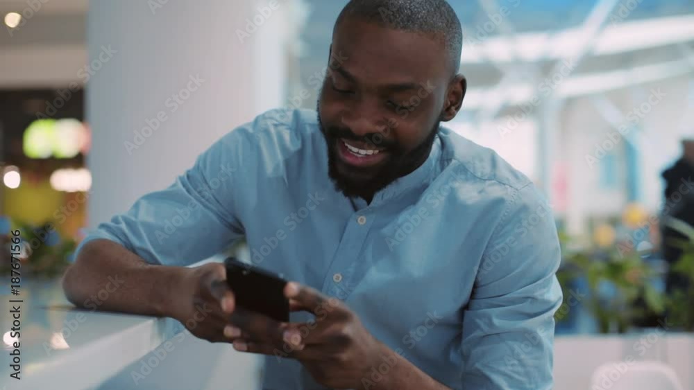 Close up face smilling african american young man using phone texting message sitting at the table in shopping center christmas internet happy black telephone portrait cute mobile holding slow motion