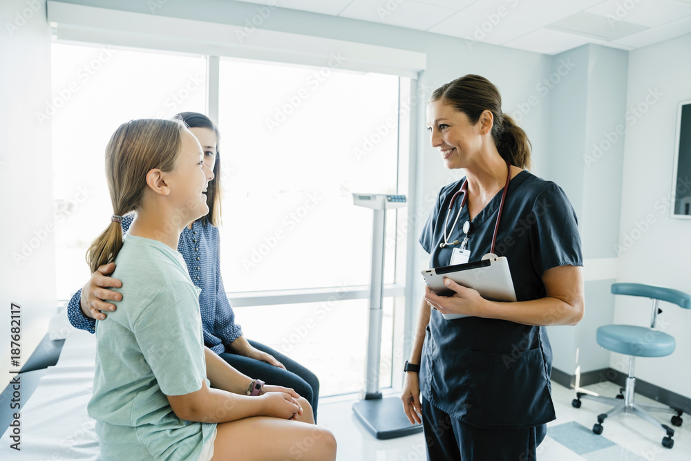 Cheerful girl looking at pediatrician while sitting by mother on examination table in hospital Cheerful girl looking at pediatrician while sitting by mother on examination table in hospital