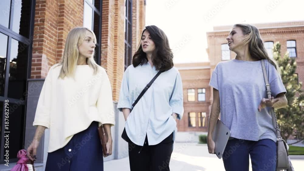 Three attractive young girls are walking in a summer street, talking and having fun. Tracking real time establishing shot