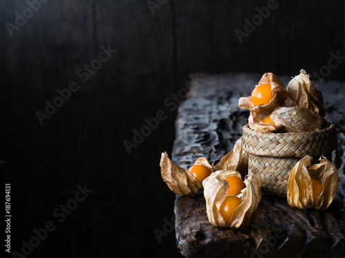 The still life of cape gooseberry in and around a small basket on a wooden slab in a dark room photo