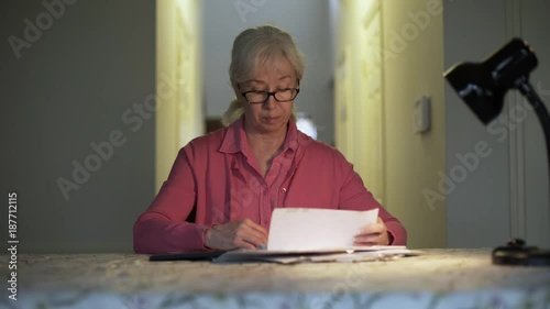 A bewildered female senior citizen on a fixed income sitting at a table who opens her check book to start paying a bill she is concerned about.