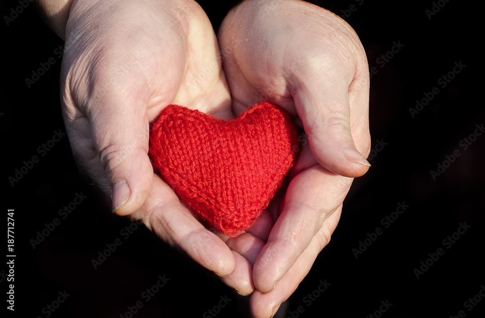 couple of hands of an elderly pensioner with wrinkled hands gently holding knitted red heart on a black background couple of hands of an elderly pensioner with wrinkled hands gently holding knitted red heart on a black background