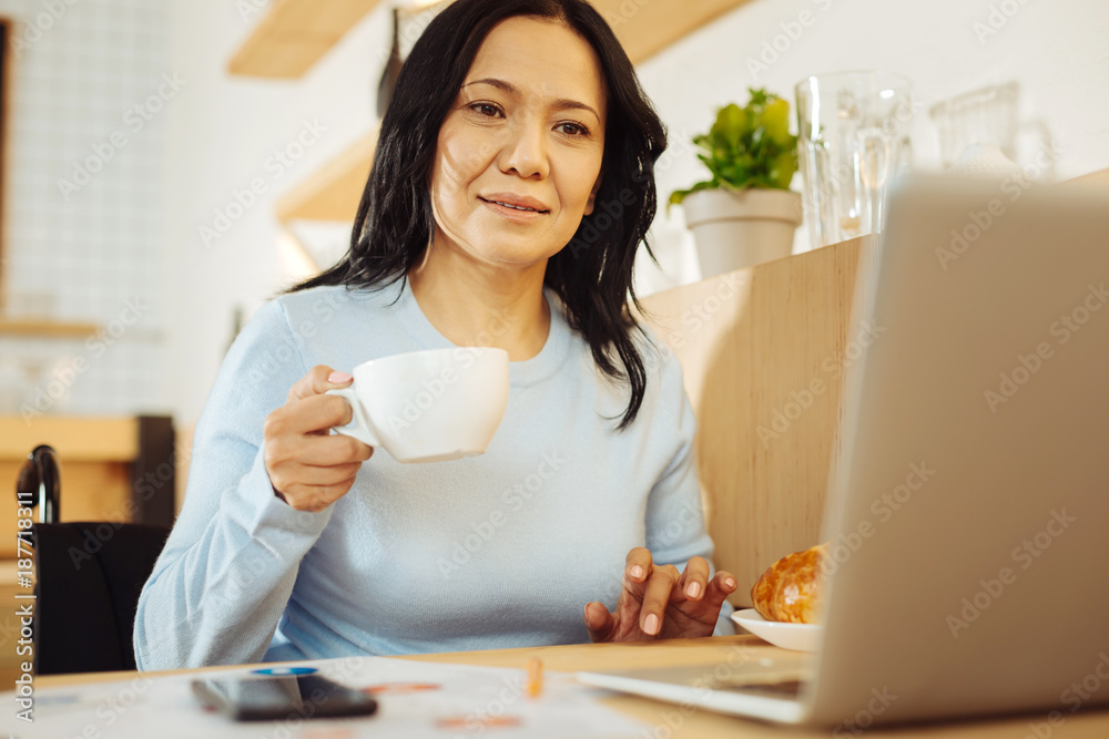 Businesswoman. Attractive alert dark-haired crippled woman sitting in a wheelchair and holding a cup of coffee and working on her laptop in a cafe Businesswoman. Attractive alert dark-haired crippled woman sitting in a wheelchair and holding a cup of coffee and working on her laptop in a cafe